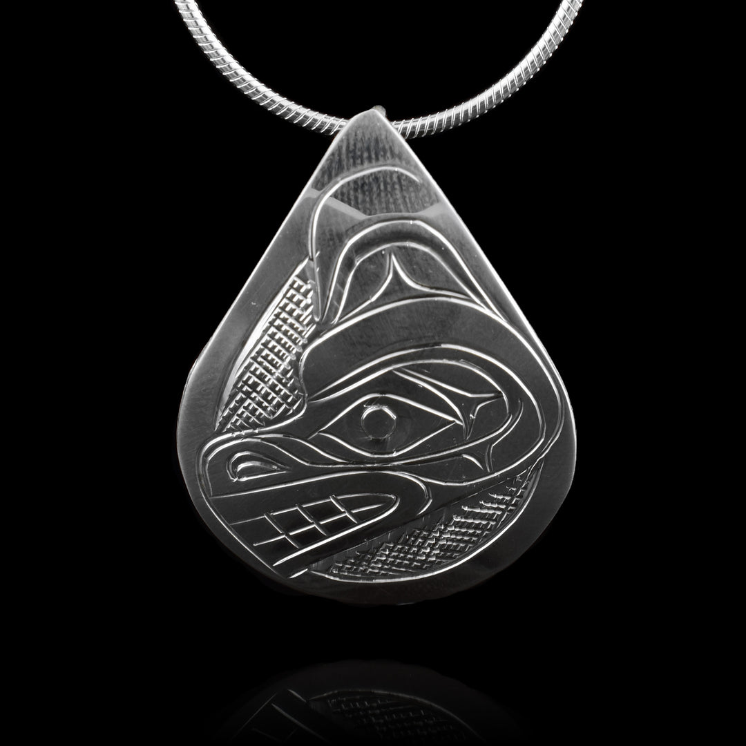 This Bear head pendant is made from sterling silver and has a large teardrop shape. There is a depiction of the head of the Bear carved in a side profile on the piece. Its snout is facing down towards the side and there are designs on its face. There are cross hatching designs in the background of the scene.
