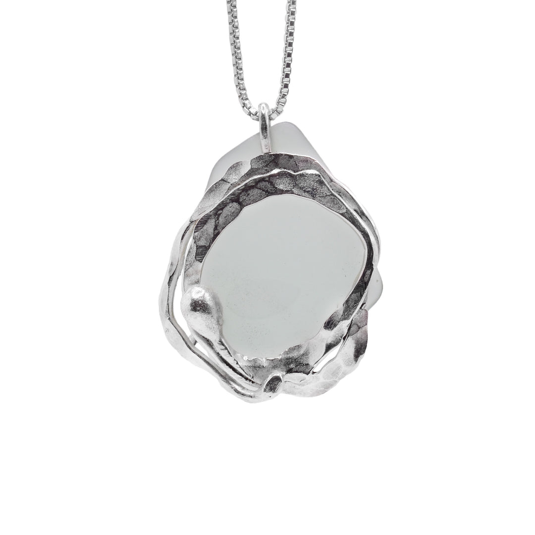 This sea glass pendant has a large, rough shaped piece and light blue sea glass dangling from a jump ring. There is a sterling silver charm in front of it that is a ring resembling water ripples.