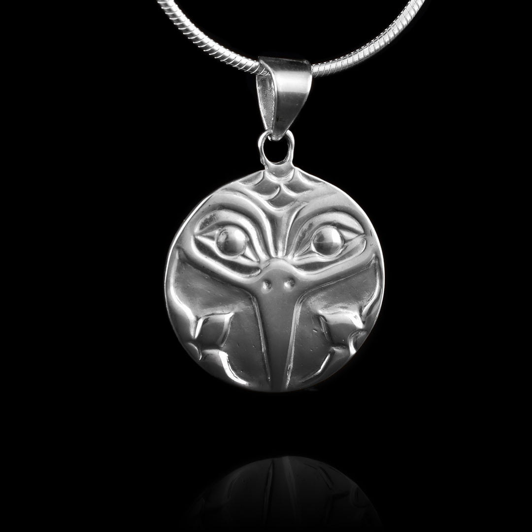 This Hummingbird pendant is made from sterling silver and is circle shaped. The piece shows the face of the Hummingbird facing out with both its eyes and thin, sharp beak detailed. There are small, flower designs at the bottom of the piece