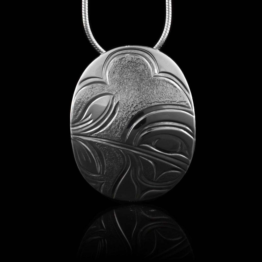 This Hummingbird pendant has a wide, oval shape and is made from sterling silver. There is a depiction of the head of the Hummingbird done at a side profile that is looking sideways and its thin beak extends across the piece into a flower. There are dimpled designs in the background of the scene.