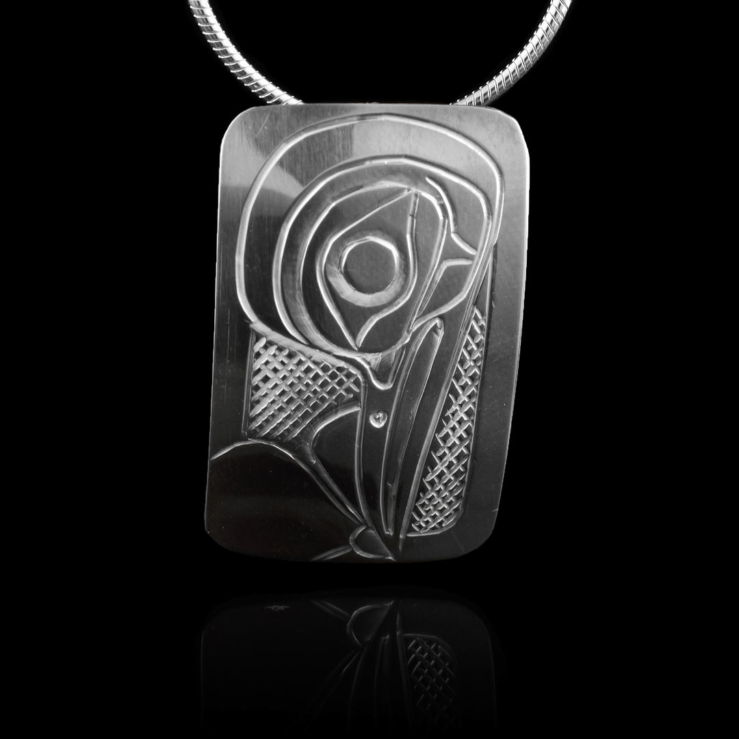 This Hummingbird head pendant is made from sterling silver and has a wide rectangle shape. There is a depiction of the head of the Hummingbird carved in a side profile on the piece. Its beak is facing down and a flower at the bottom of the piece. There are cross hatching designs in the background of the scene.