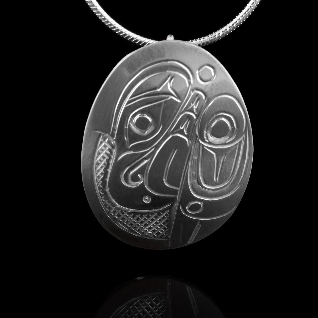 This Eagle head pendant is made from sterling silver and has an oval shape. There is a depiction of the head of the Eagle and its wing carved in a side profile on the piece. Its beak is facing down and its wing has designs on it and runs below the beak. There are cross hatching designs in the background of the scene.