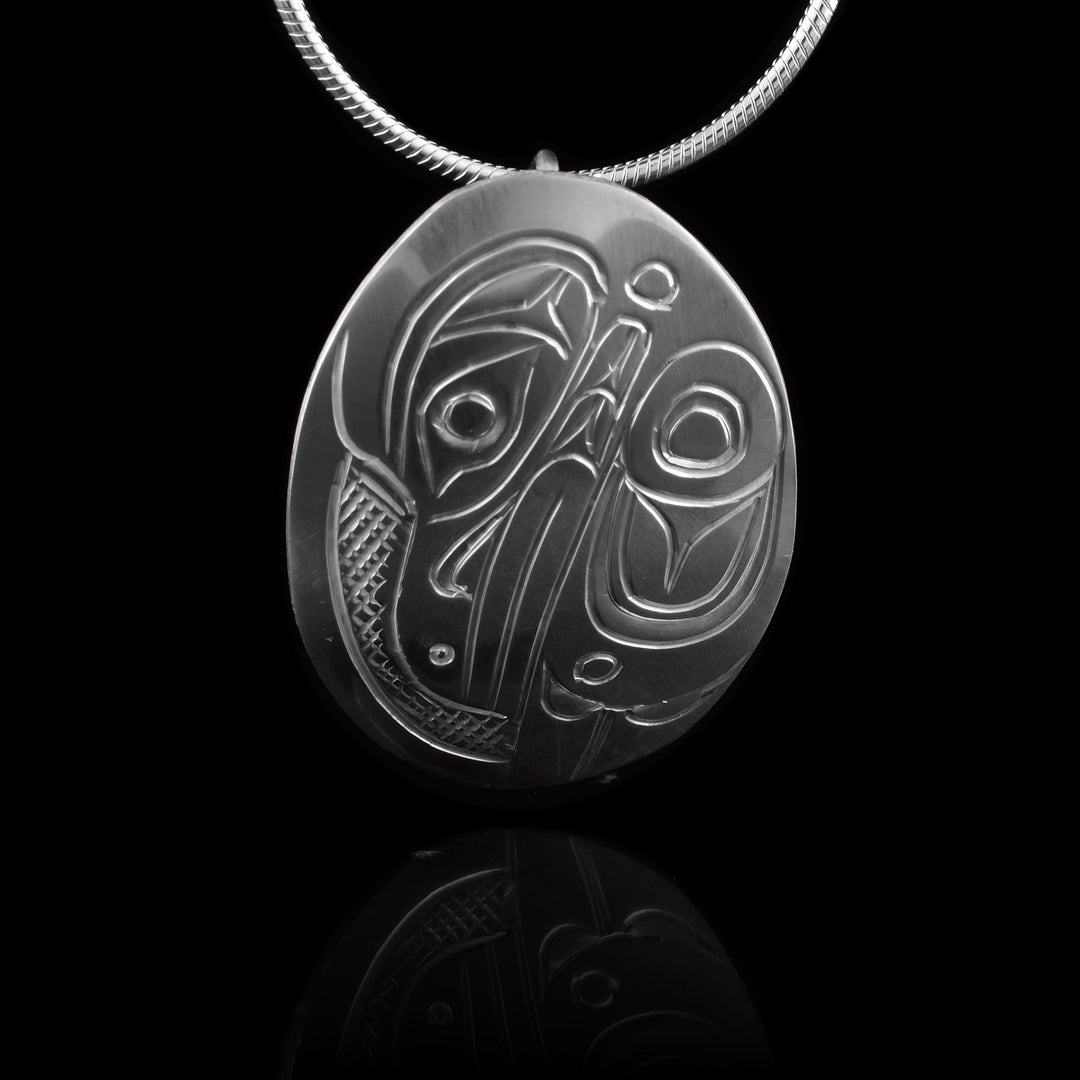 This Raven pendant is made from sterling silver and has a small oval shape. There is a depiction of the head and wing of the Raven carved in a side profile on the piece. Its beak is facing down and its wing runs along the back of the beak. There are cross hatching designs in the background of the scene.