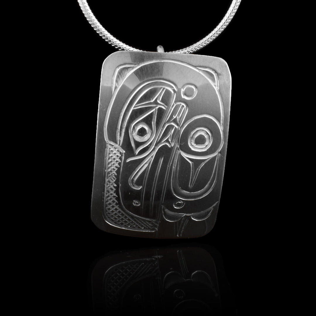 This Raven pendant is made from sterling silver and has a small rectangle shape. There is a depiction of the head and wing of the Raven carved in a side profile on the piece. Its beak is facing down and its wing runs along the back of the beak. There are cross hatching designs in the background of the scene.