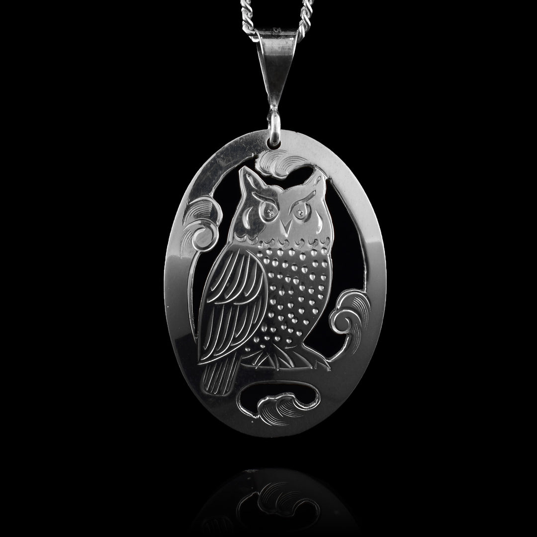 This sterling silver owl pendant has an oval shape and an owl is depicted in the  center of it. There are cutouts around the owl on a branch and carvings on the surface of the piece depicting the bird. There are wind designs around it.