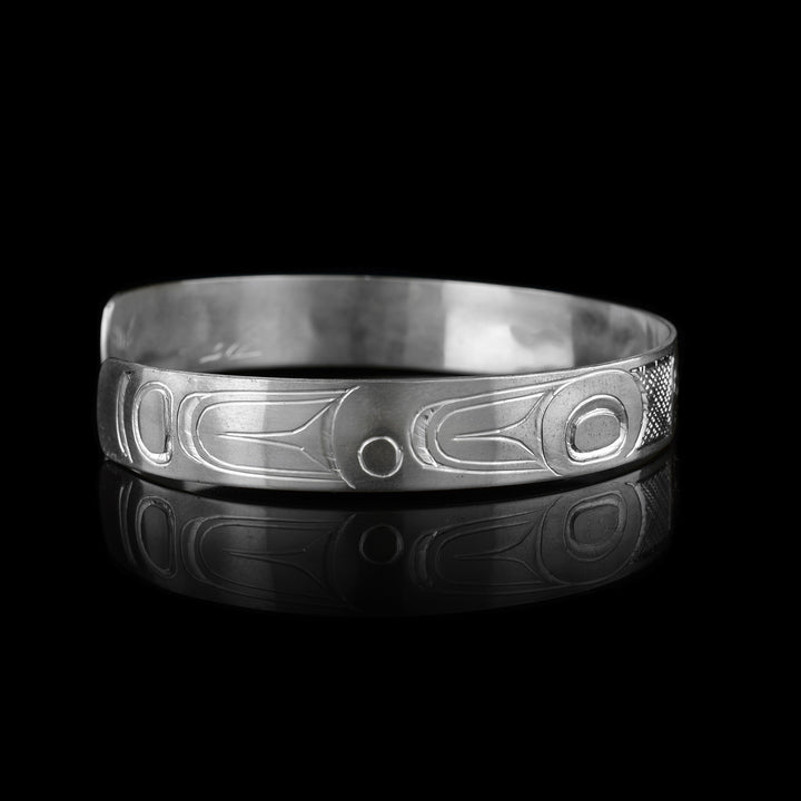 This dainty Orca bracelet has a thin, sterling silver band with a gap in the back. The head of the Orca is carved in the center of the band and takes up the width of the band. The head has an oval shape, is at a side profile and its facing the left. On both ends, there are circular, oblong and triangular patterns depicting the rest of the legend. There are cross hatching designs in the background of this piece.