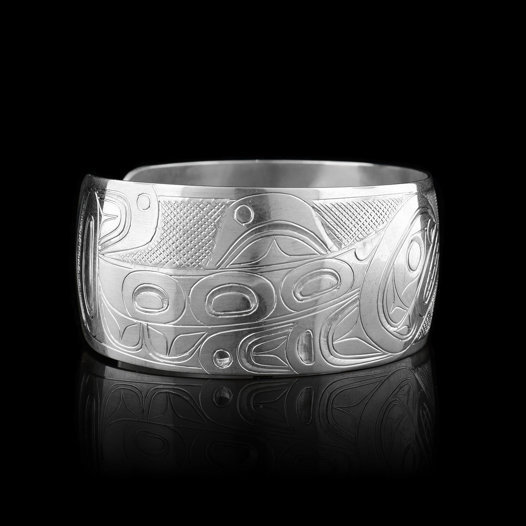 This Salmon and Orca bracelet has a wide, sterling silver band with a gap in the back. The body of the entire Salmon is depicted in a side profile on the left of the piece. This legend has a signature hump on its back and its head is facing upwards in the center of the piece. The entire Orca is depicted on the right. It has its dorsal fin and its head is breaching downwards in the center of the band. There are cross hatching patterns in the background of the piece.