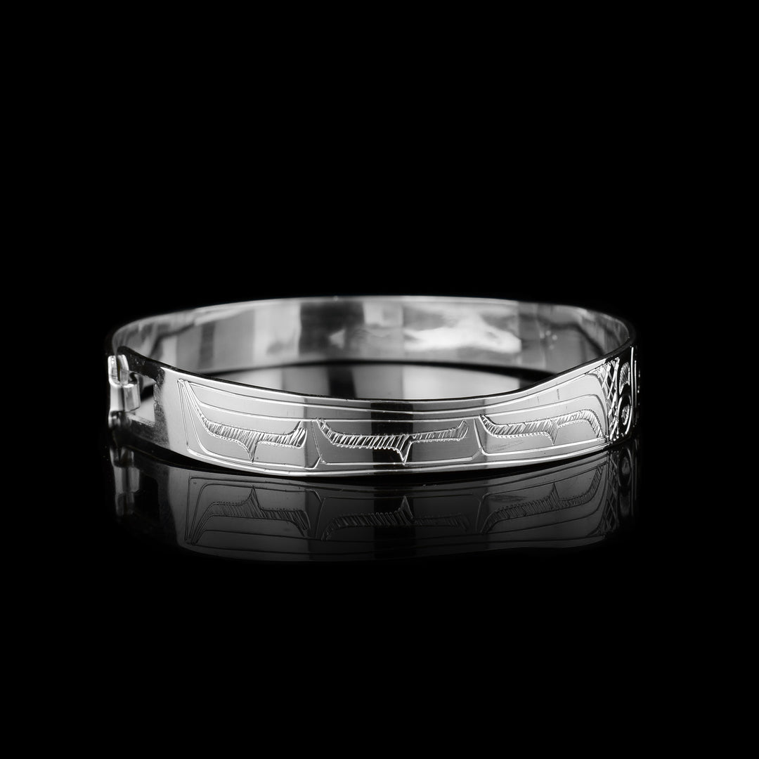 This dainty bracelet has a thin, sterling silver band with a clasp in the back. There is are depictions two Hummingbirds facing each other carved into the surface of the band. Their wings are carved behind the heads and have circular and triangular designs carved into them. There are cross hatching patterns in the background of this piece.