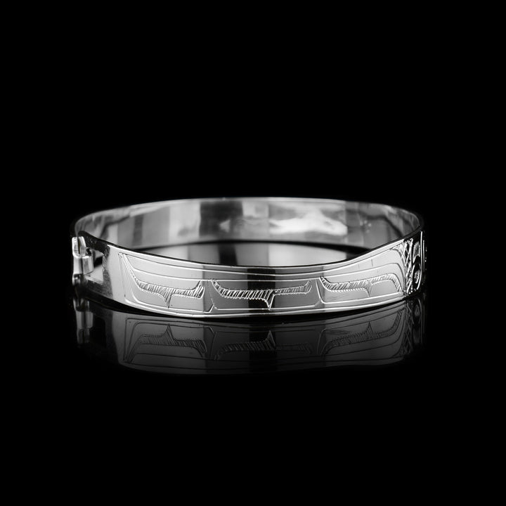 This dainty bracelet has a thin, sterling silver band with a clasp in the back. There is are depictions two Hummingbirds facing each other carved into the surface of the band. Their wings are carved behind the heads and have circular and triangular designs carved into them. There are cross hatching patterns in the background of this piece.