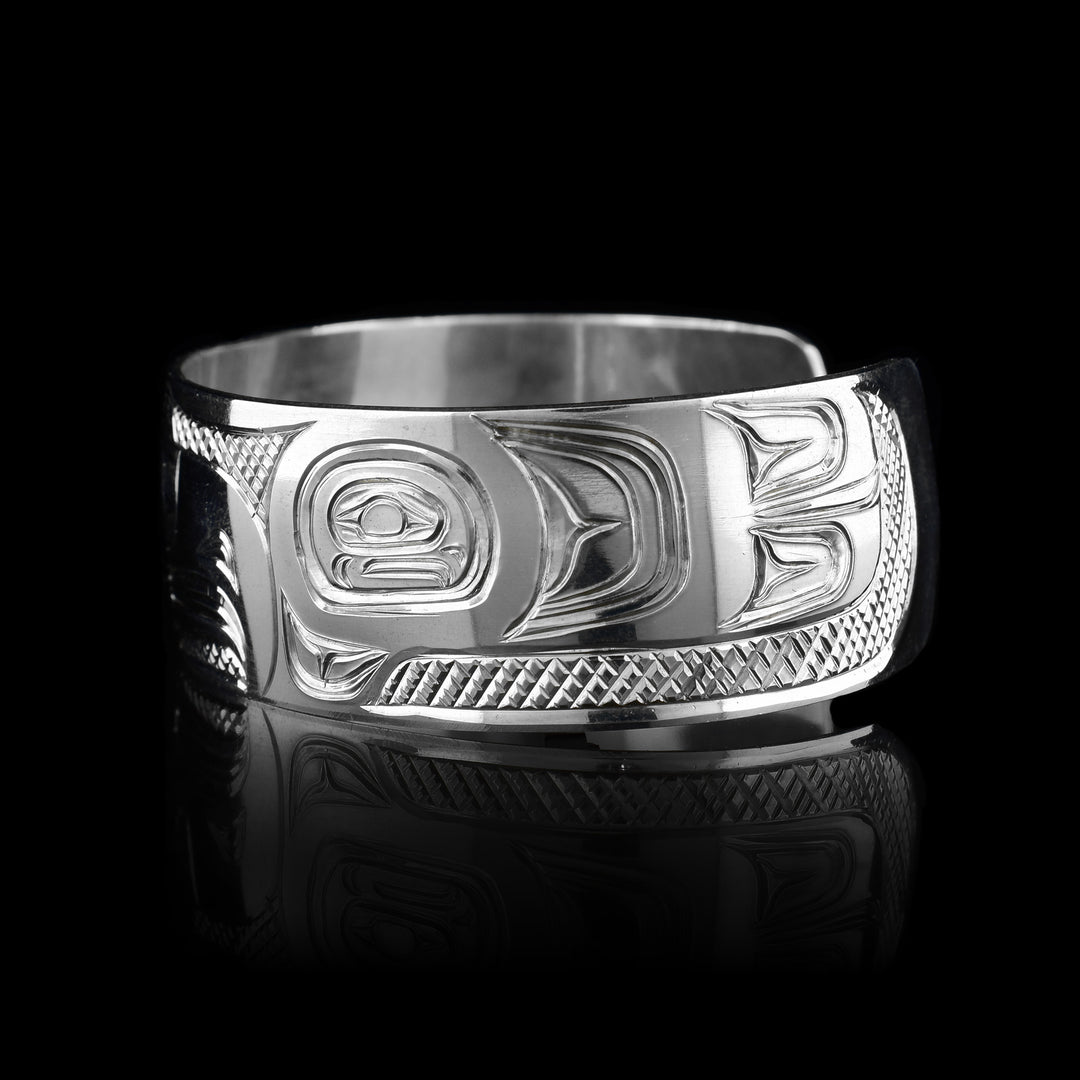 This Thunderbird bracelet cuff has been made using sterling silver and has a single, wide band with a gap in the back. The Thunderbird's head is carved into the center of the piece in a side profile and takes up the width of the band. Its two wings are depicted around it on the rest of the band. There are feather details made into the wings and the head as well as cross hatching patterns in the background of this piece.
