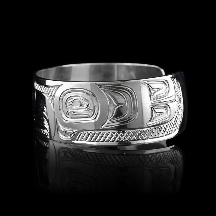 This Thunderbird bracelet cuff has been made using sterling silver and has a single, wide band with a gap in the back. The Thunderbird's head is carved into the center of the piece in a side profile and takes up the width of the band. Its two wings are depicted around it on the rest of the band. There are feather details made into the wings and the head as well as cross hatching patterns in the background of this piece.