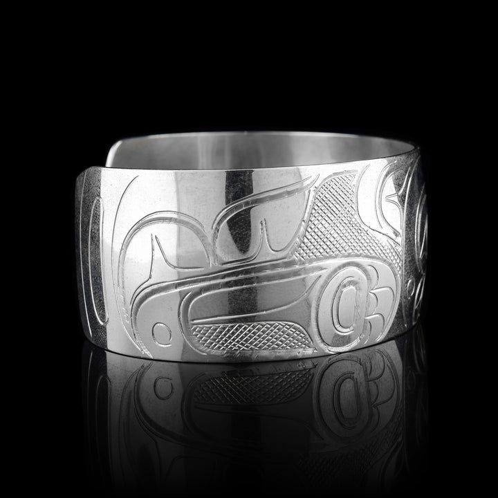 This Wolf bracelet has a wide, sterling silver band with a gap in the back. The head of the Wolf is carved in the center of the piece in a side profile and takes up the width of the band. To the right, there is one of its clawed paws and its tail curling up to the top of the band. On the left its other leg is shown.
