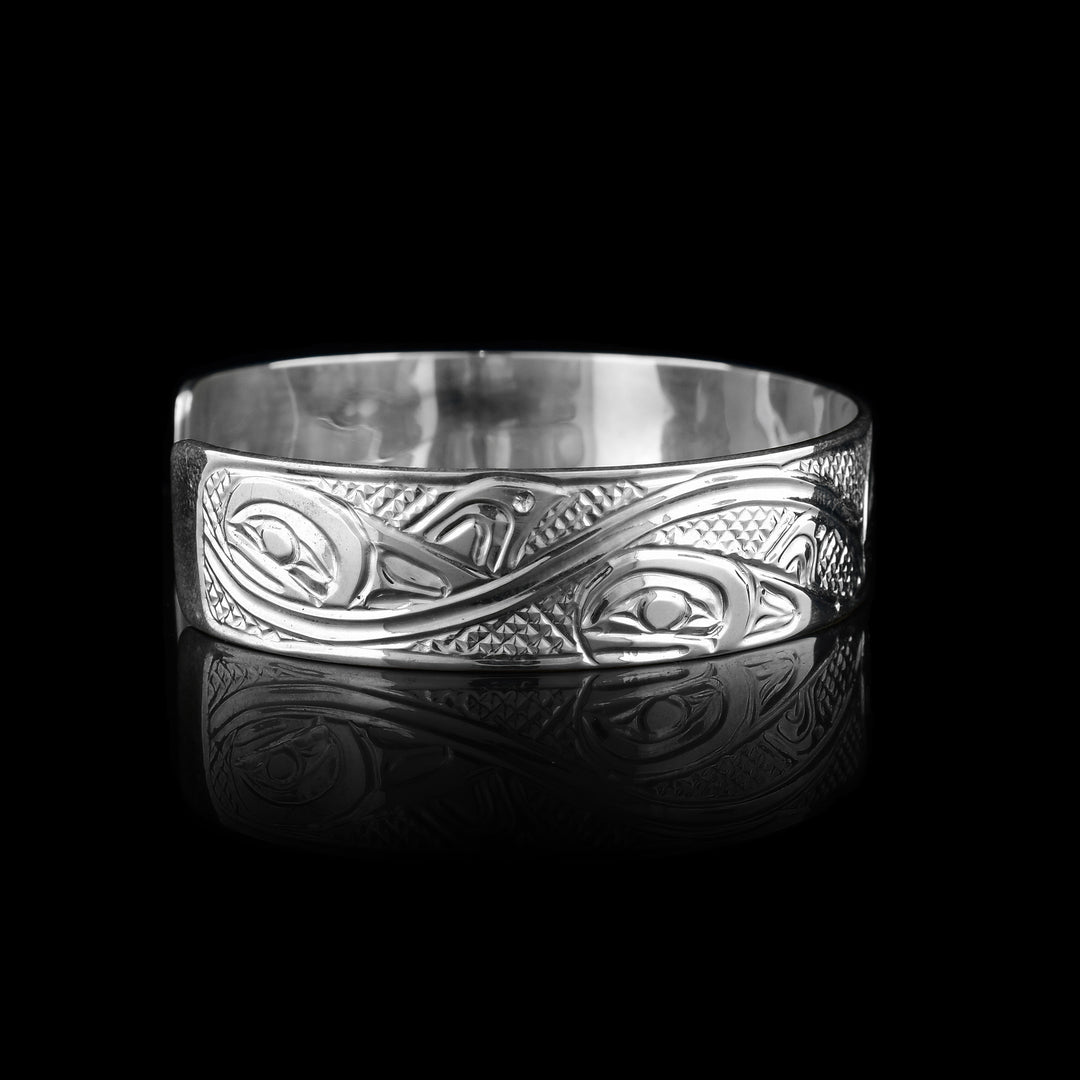 This sterling silver cuff bracelet has a single band with a gap in the back and a depiction of the Orca legend carved into the surface of it. On the right, there are two Orca's depicted in a side profile with large heads and their dorsal fins in view. On the left, there are wave designs with smaller whales breaching from them representing an Orca pod.