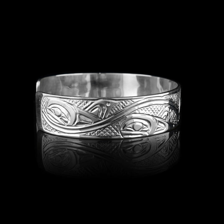This sterling silver cuff bracelet has a single band with a gap in the back and a depiction of the Orca legend carved into the surface of it. On the right, there are two Orca's depicted in a side profile with large heads and their dorsal fins in view. On the left, there are wave designs with smaller whales breaching from them representing an Orca pod.