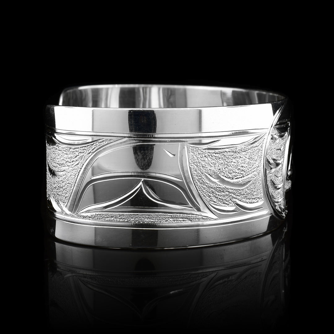 This stunning Orca bracelet is made using sterling silver and has a single, wide band with a gap in the back. There are intricate carvings on the surface of the piece that depicts the Baby Orca in the womb and being watched over by its mother who is carved on the side of the piece. On the other side of the band, there is a dorsal fin in the sea depicted.