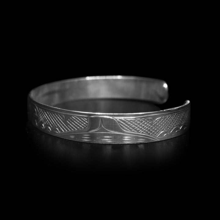 This dainty bracelet has a thin, sterling silver band with a gap in the back. The head of the Orca is carved in the center of the band and there are the backs of other whales with their dorsal fins breaching out of wave patterns. There are cross hatching patterns in the space behind the legends.