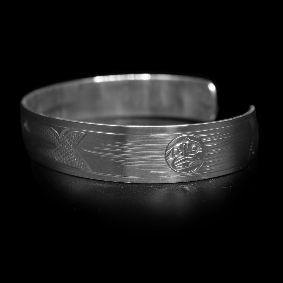 This Hummingbird bracelet has a single, sterling silver band with a gap in the back. There is a depiction of the Hummingbird carved across the surface of the band and a small Sun carved in the side of the band. The Hummingbird is depicted in a side profile and is facing the Sun. The head of the legend is in the center of the band and there are cross hatching designs in the negative space of the piece.