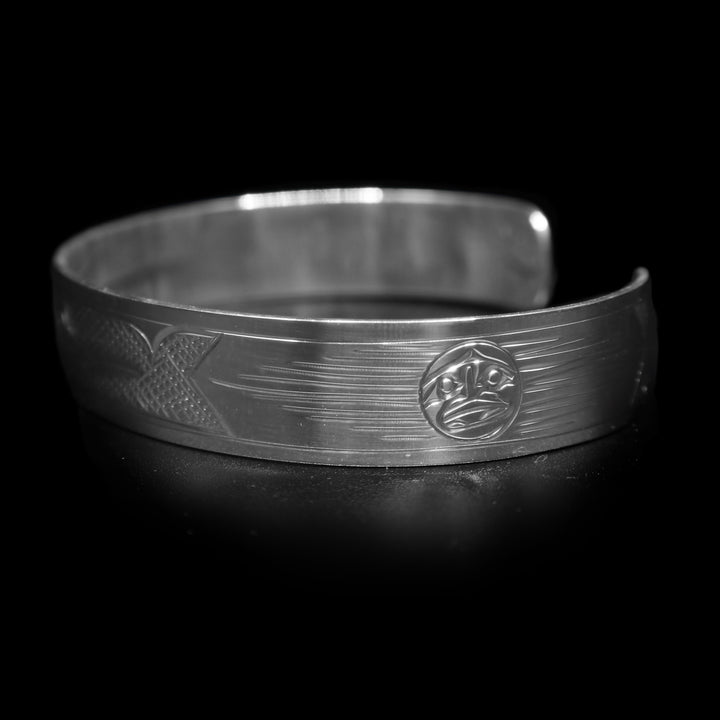 This Hummingbird bracelet has a single, sterling silver band with a gap in the back. There is a depiction of the Hummingbird carved across the surface of the band and a small Sun carved in the side of the band. The Hummingbird is depicted in a side profile and is facing the Sun. The head of the legend is in the center of the band and there are cross hatching designs in the negative space of the piece.