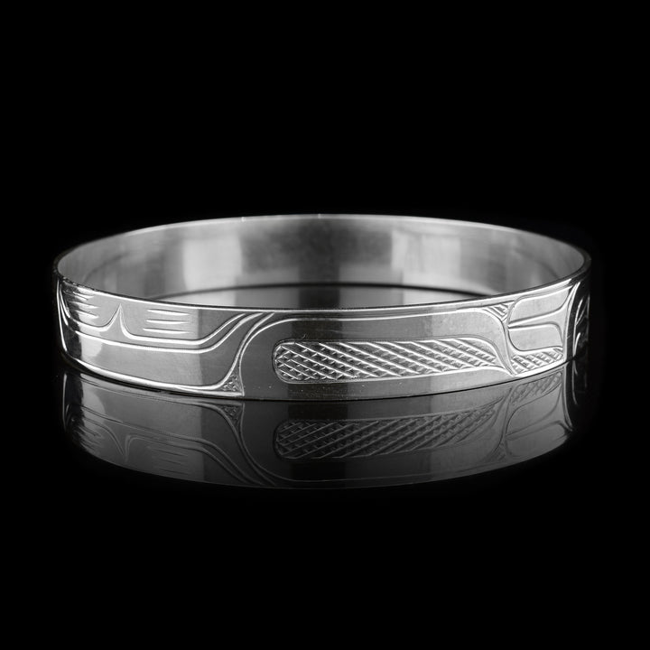 This sterling silver bangle bracelet has a single, thin band that wraps around the wrist. There are carvings across the surface of the band depicting the Raven.