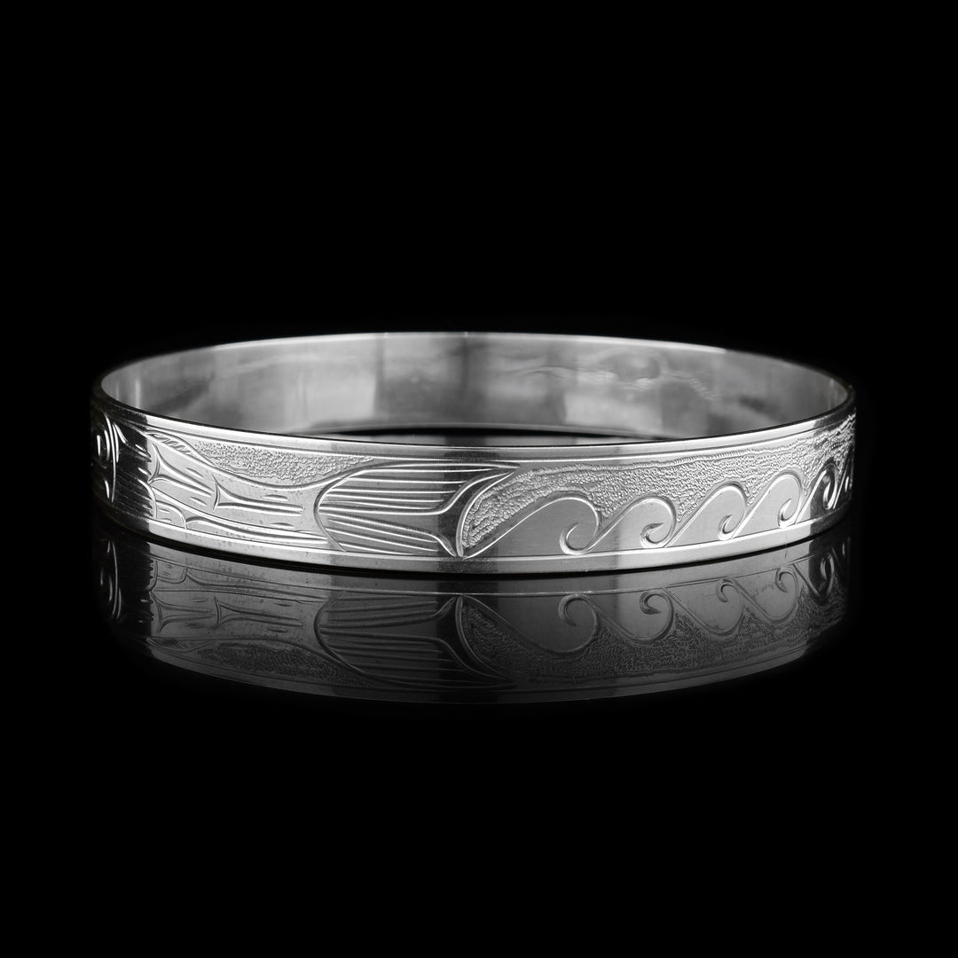 This Salmon and Orca bracelet bangle is made from sterling silver and has a single, thin band. There are depictions of the Salmon and the Orca carved across the surface of the band.