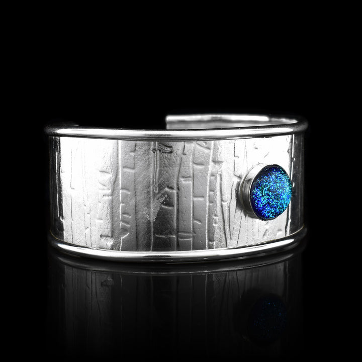 This wide bracelet cuff has has a single, wide band made from sterling silver and has a gap in the back. The edges of the band are raised and there are striped and abstract designs that have been pressed into the surface of it. There is a light blue colored fused glass bead that is embossed into the top of the band.