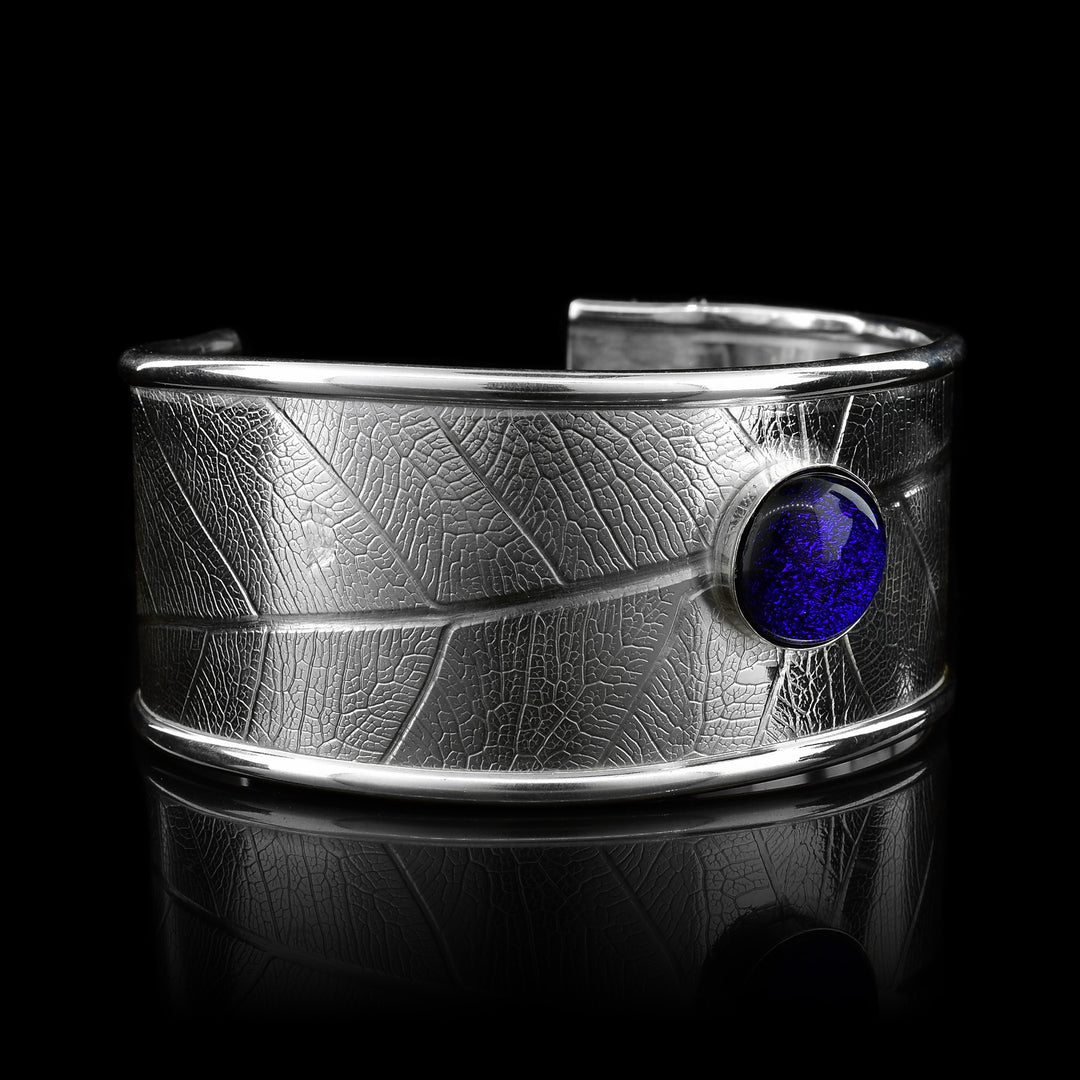 This beautiful leaf pattern, wide cuff bracelet is made using sterling silver and a royal blue colored fused glass bead. The piece has a single, wide band with a gap in the back and there are detailed leaf textures imprinted across the surface of it. The glass bead has interesting details on the surface of it and is embossed in the top, center of the bracelet
