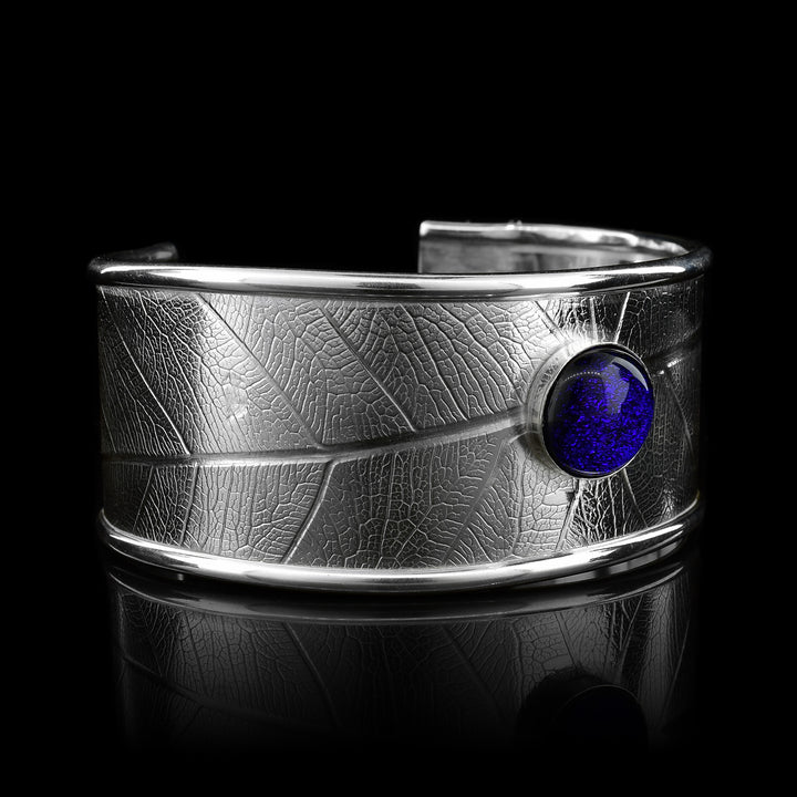 This beautiful leaf pattern, wide cuff bracelet is made using sterling silver and a royal blue colored fused glass bead. The piece has a single, wide band with a gap in the back and there are detailed leaf textures imprinted across the surface of it. The glass bead has interesting details on the surface of it and is embossed in the top, center of the bracelet