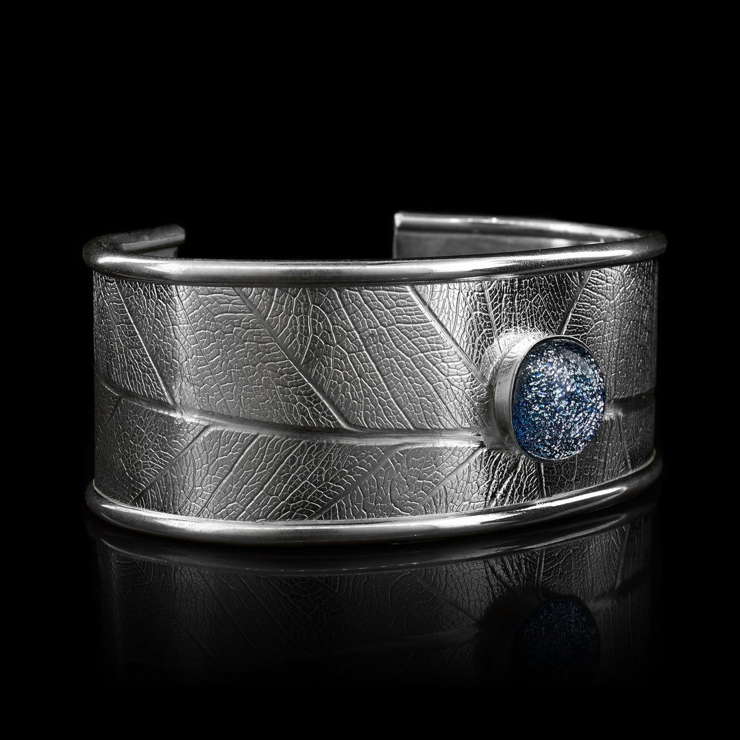 This beautiful leaf pattern, wide cuff bracelet is made using sterling silver and a denim (blue and white speckled) colored fused glass bead. The piece has a single, wide band with a gap in the back and there are detailed leaf textures imprinted across the surface of it. The glass bead has interesting details on the surface of it and is embossed in the top, center of the bracelet