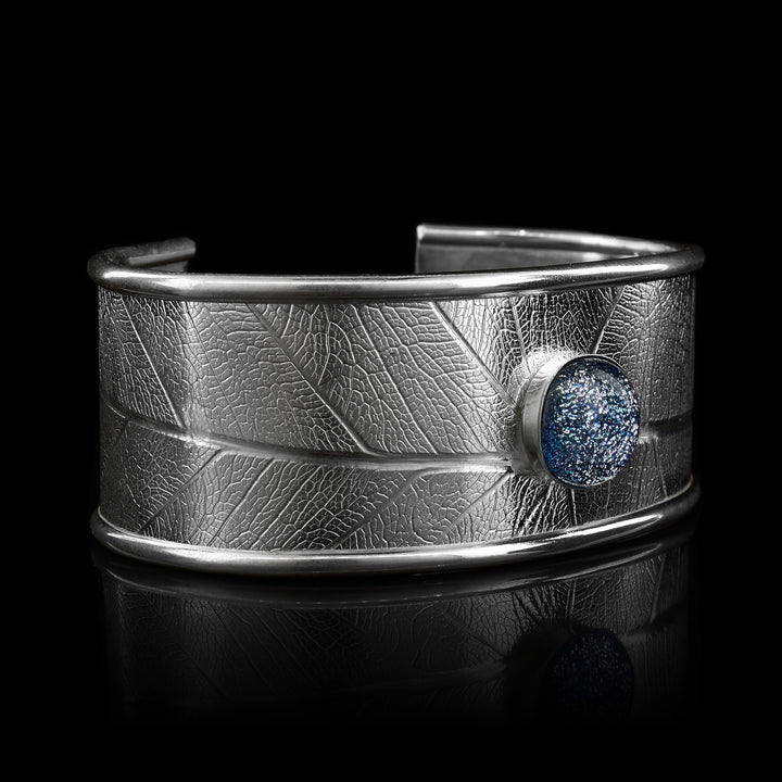 This beautiful leaf pattern, wide cuff bracelet is made using sterling silver and a denim (blue and white speckled) colored fused glass bead. The piece has a single, wide band with a gap in the back and there are detailed leaf textures imprinted across the surface of it. The glass bead has interesting details on the surface of it and is embossed in the top, center of the bracelet