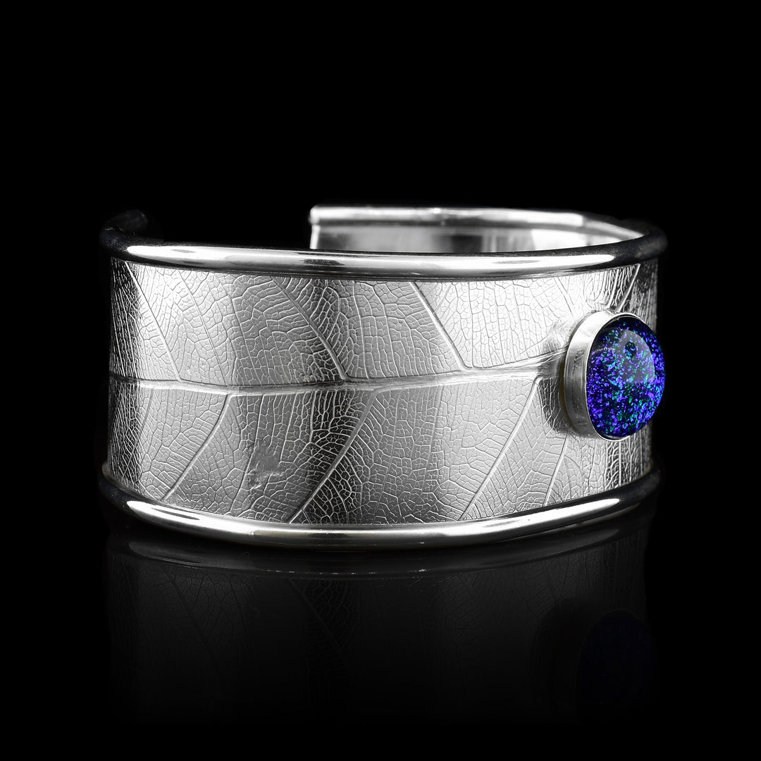 This beautiful leaf cuff bracelet is made using sterling silver and a turquoise colored fused glass bead. The piece has a single, wide band with a gap in the back and there are detailed leaf textures imprinted across the surface of it. The glass bead has interesting details on the surface of it and is embossed in the top, center of the bracelet