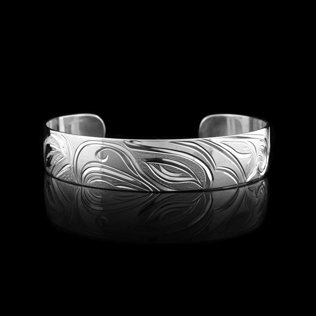 This Salmon and Thunderbird bracelet cuff has a single, sterling silver band with a gap in the back. The head of the Thunderbird is done at a side profile in the center of the band with a salmon depicted on both ends of the band.