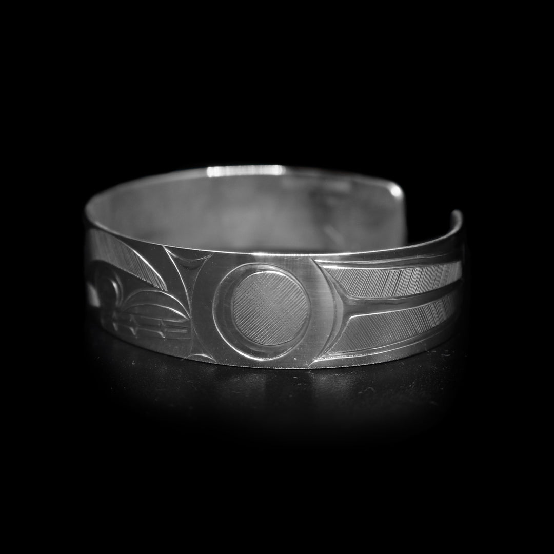 This Orca whale bracelet has a single, sterling silver band with a gap in the back. There is a depiction of the Orca legend intricately carved into the surface of the band. The head of the legend is in the center of the band and its fins are depicted on either side of the head. The accents are made with fine, hatching patterns.
