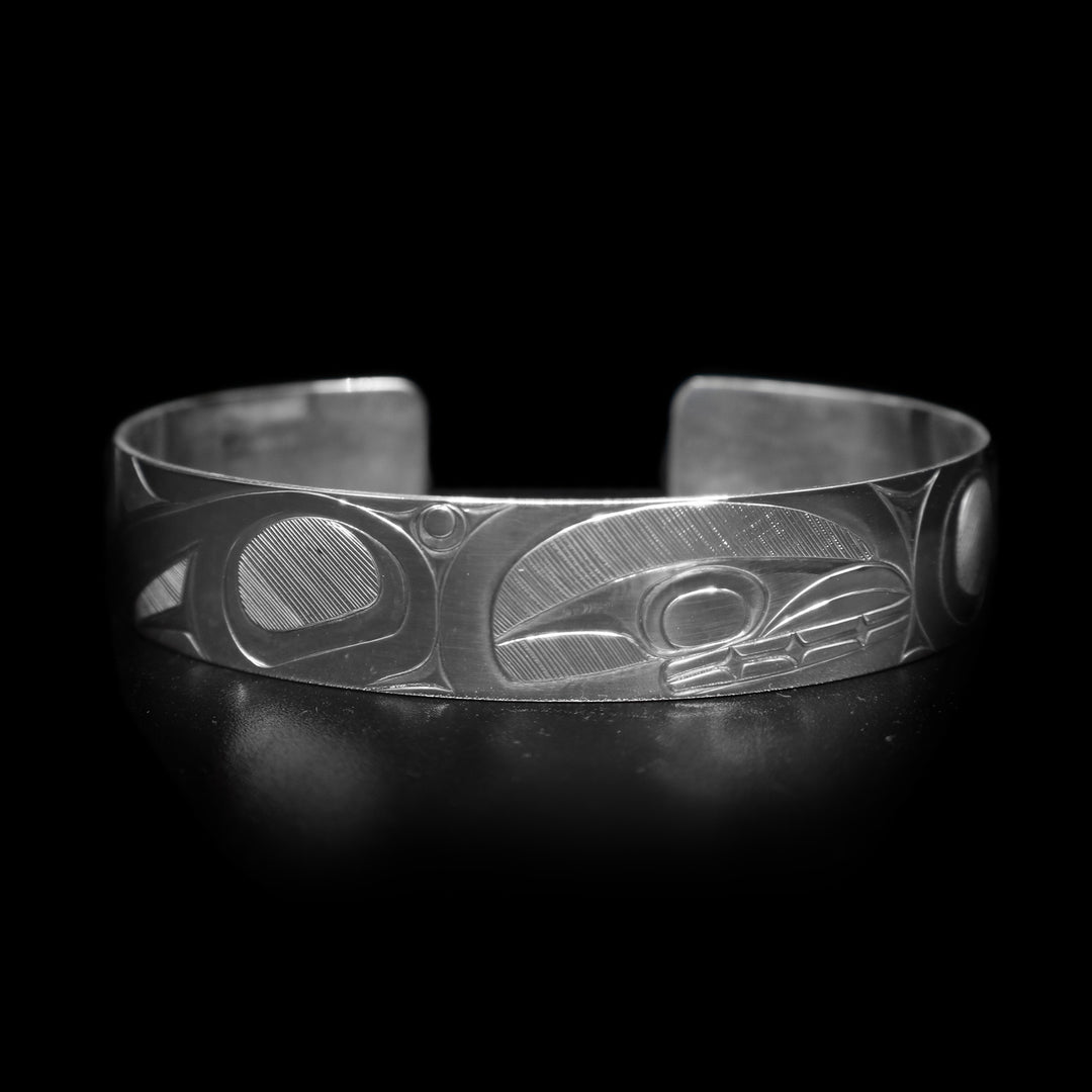 This Orca whale bracelet has a single, sterling silver band with a gap in the back. There is a depiction of the Orca legend intricately carved into the surface of the band. The head of the legend is in the center of the band and its fins are depicted on either side of the head. The accents are made with fine, hatching patterns.