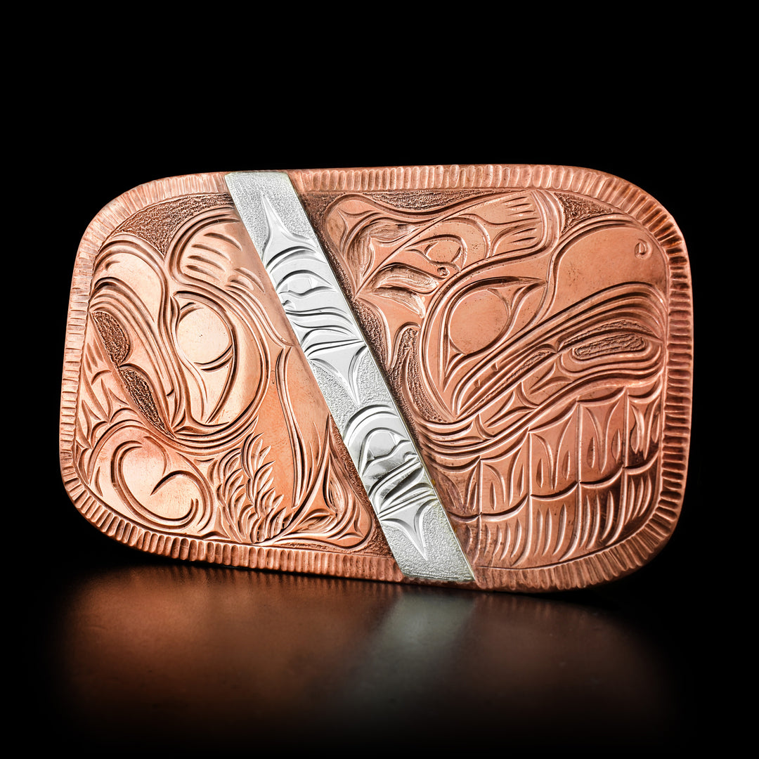This copper eagle belt buckle depicts two eagles facing away from each other on each side of the buckle. In the center is a lopsided piece of silver with carvings on it.