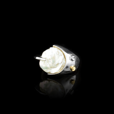 This pearl signet ring has a thin, sterling silver band and a large, flat pearl that acts as the signet for the piece. The silver band widens at the top and wraps around the circumference of the pearl. There is a 14K gold stud on the wide part of the band.