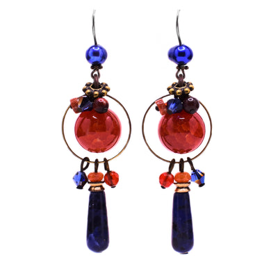 These dangle earrings feature a large, round carnelian agate in the center with small gemstones on top as accents. Around it is a thin brass ring with a teardrop sodalite and more gemstones attached at the bottom. At the top of each earring is an assymetrical blue pearl.