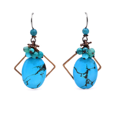 These turquoise earrings feature a light blue, oval piece of turquoise hanging from a cluster of blue gemstones with a diamond-shaped brass accent behind it.