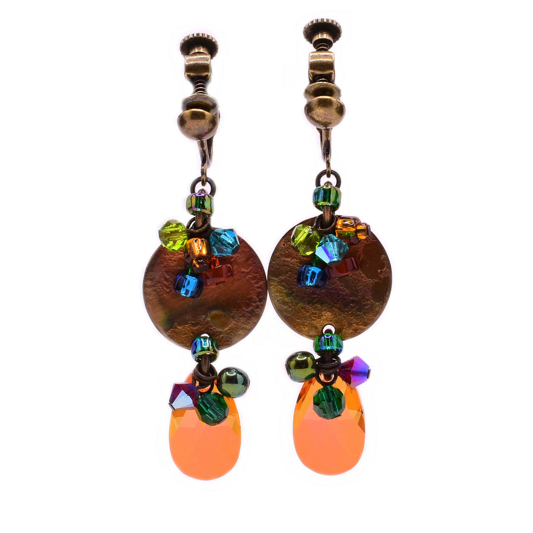 These clip earrings feature a circular brass accent in the center with a cluster of multi coloured gemstones hanging from the top. At the bottom of the brass accent a teardrop orange crystal is attached with another cluster of multi coloured gemstones.