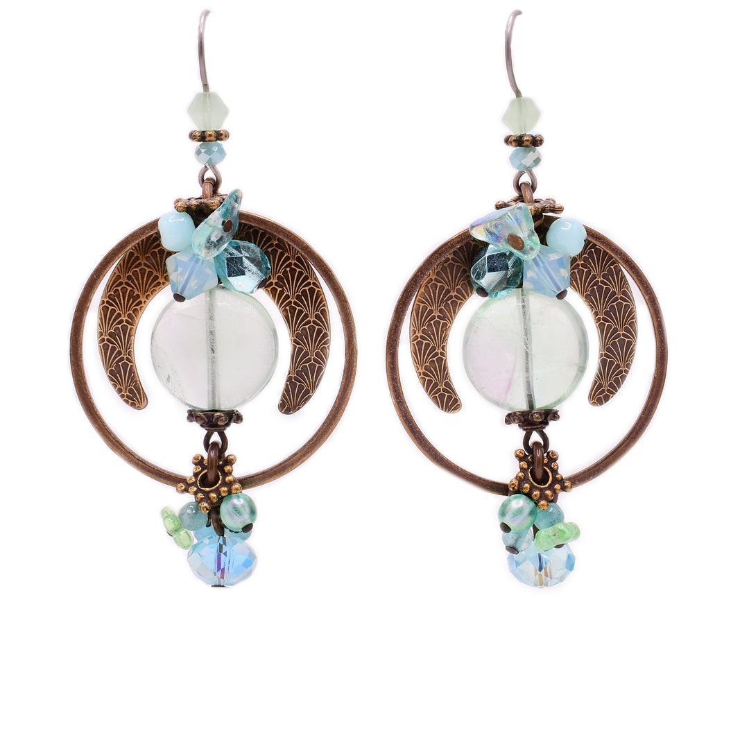 These glass earrings feature a round glass piece in the center with a uniquely shaped brass accent above and a large, circular brass accent around it. Hanging below the glass is a cluster of light blue and green gemstones. Above the glass piece is another cluster with similar gemstones.