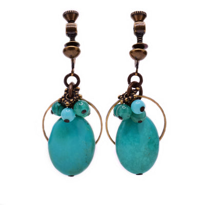These turquoise earrings feature an oval turquoise as the centerpiece with a cluster of light blue and green gemstones above. There is a thin, circular brass accent behind the turquoise and additional brass accents on top of the cluster.