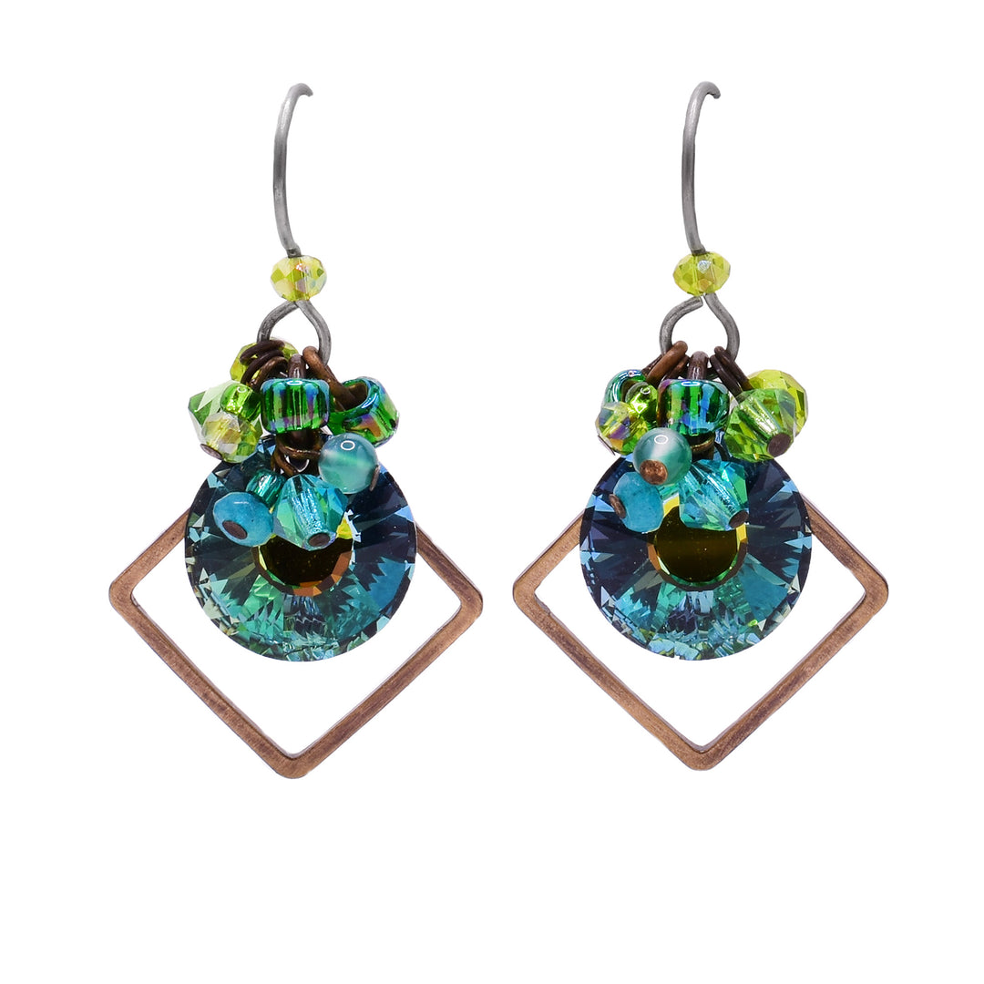These Round Dangle Earrings feature a circular, blue glass in the center with a cluster of light green and blue gemstones above. There is a thin, diamond-shaped brass accent behind. A light green crystal has been placed right underneath the titanium hook.