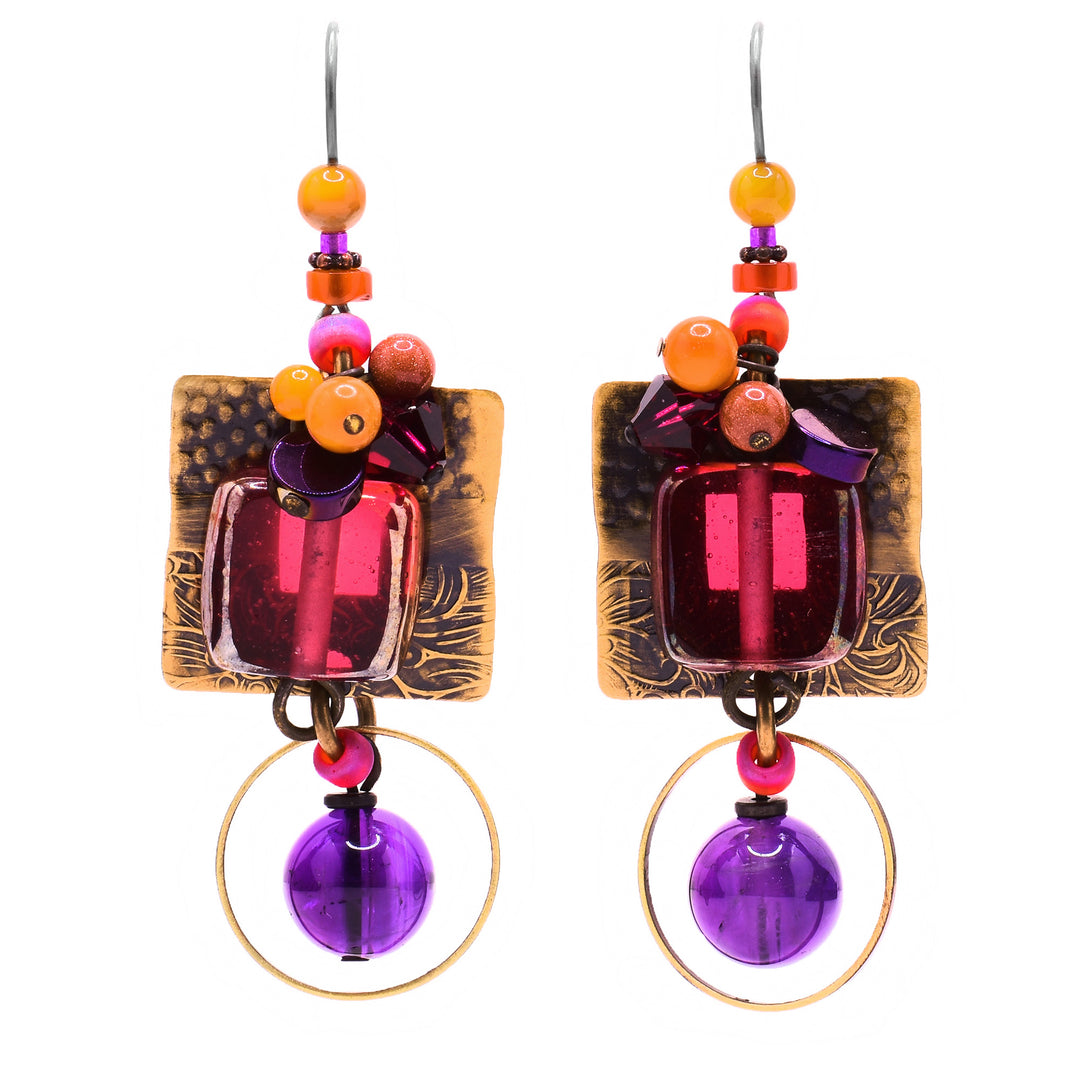 These glass earrings feature a square pink glass in the center with a cluster of orange, purple, and pink gemstones on top. Behind is a square brass accent with a floral and polka dot design. Attached to the bottom is a round amethyst bead with a circular brass accent.