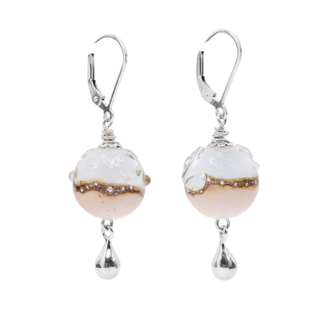 These ocean earrings have sphere shaped, glass hangs that dangle from sterling silver. The top half of the ball is blue and textured like water, the bottom is a light color meant to resemble sand. Going around the equator of the balls is a dark band with dots in it that is wavy. There are silver teardrops dangling below th glass balls.
