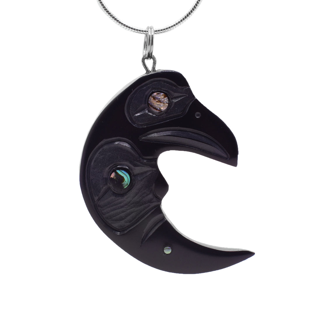 This Raven Moon pendant has been carved from argillite stone and has the shape of a crescent moon. At the top of the crescent, there is the head of the Raven with an abalone gem in its eye. The moon legend has a large piece of abalone as its eye and there are carvings across it depicting the legends. 