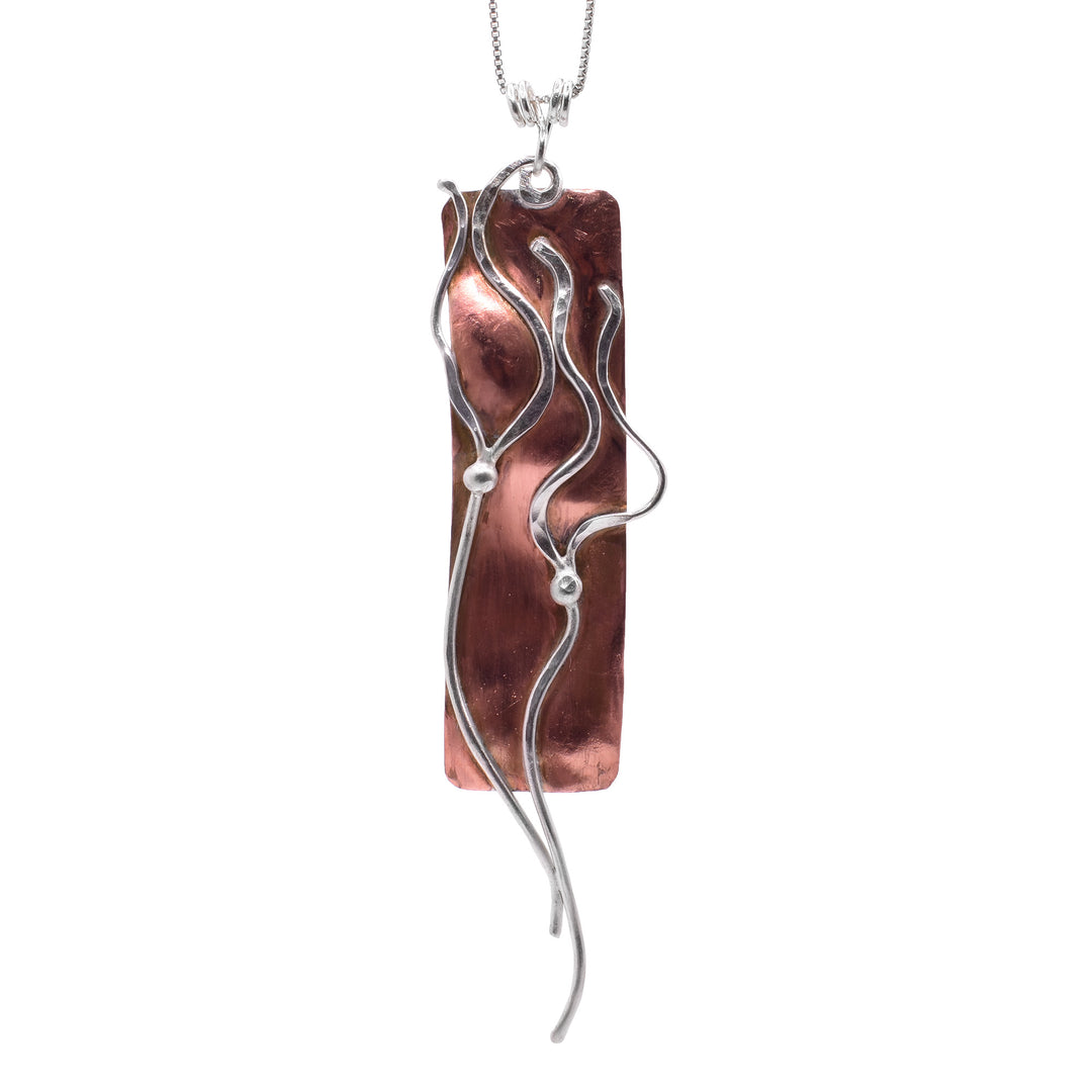 This ocean pendant necklace has a long, thin rectangle made from copper with sterling silver charms on the surface of it. The silver is shaped like bull kelp and it extends past the copper part of the pendant.