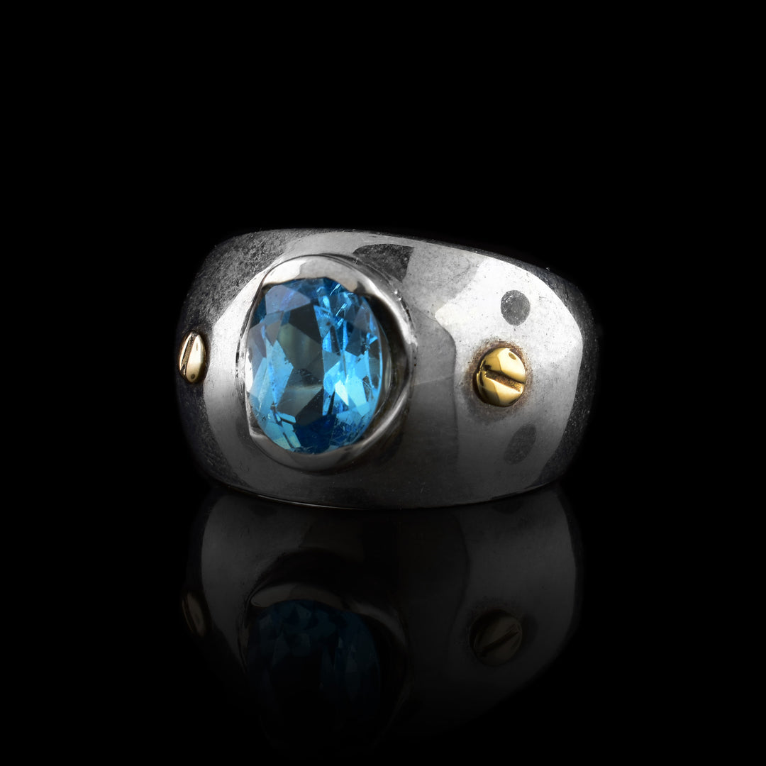 These blue topaz rings have wide, tapered and sterling silver bands with a large oval shaped blue topaz gem embossed in the center of the solitaire ring. There are two 14K gold studs placed on either side of the gem.