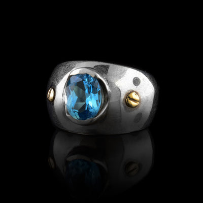 These blue topaz rings have wide, tapered and sterling silver bands with a large oval shaped blue topaz gem embossed in the center of the solitaire ring. There are two 14K gold studs placed on either side of the gem.