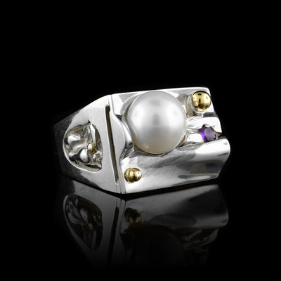 This large pearl signet ring has a wide, sterling silver band that tapers slightly. The top of the band is a signet with a wavy surface and there is a large pearl, two gold studs and an amethyst on the surface of the signet. These pieces are nestled in the waves and on the side there are wave designs.