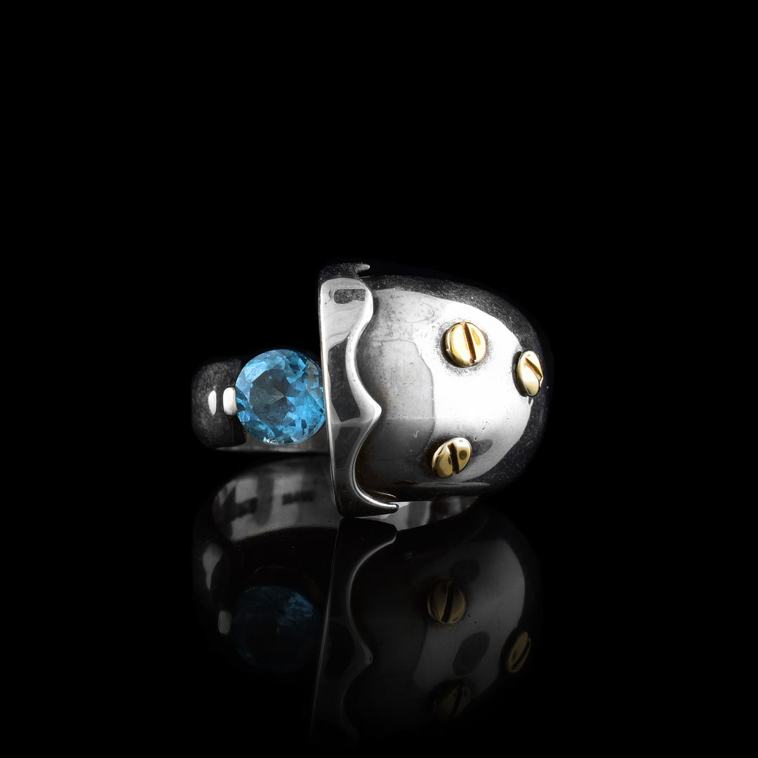 This sterling silver topaz rings has an abstract shape with one end of the band being very large and round with designs on it. There are 14K gold studs on the large part of the ring and a blue topaz gem held between the band.