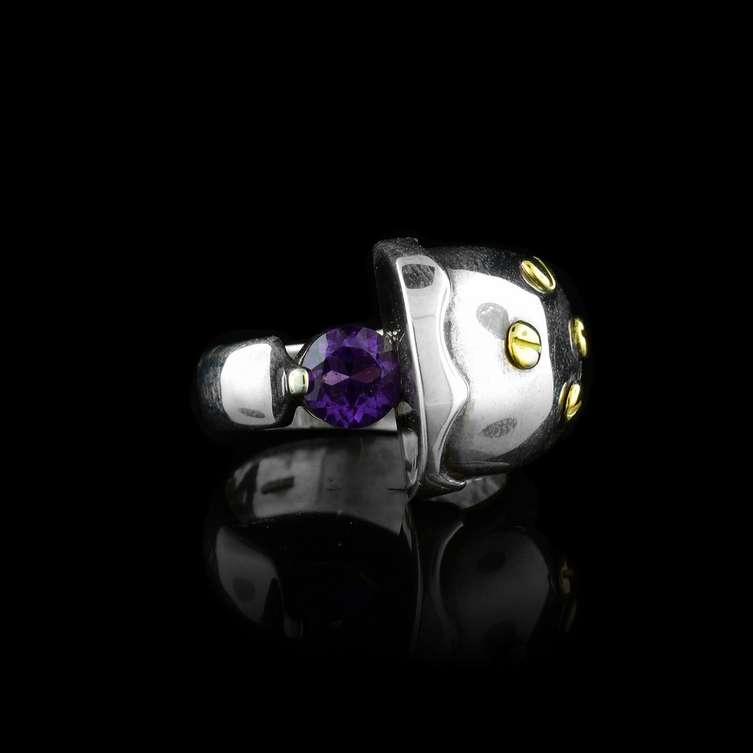 This sterling silver amethyst ring has an abstract shape with one end of the band being very large and round with designs on it. There are 14K gold studs on the large part of the ring and an amethyst gem held between the band.